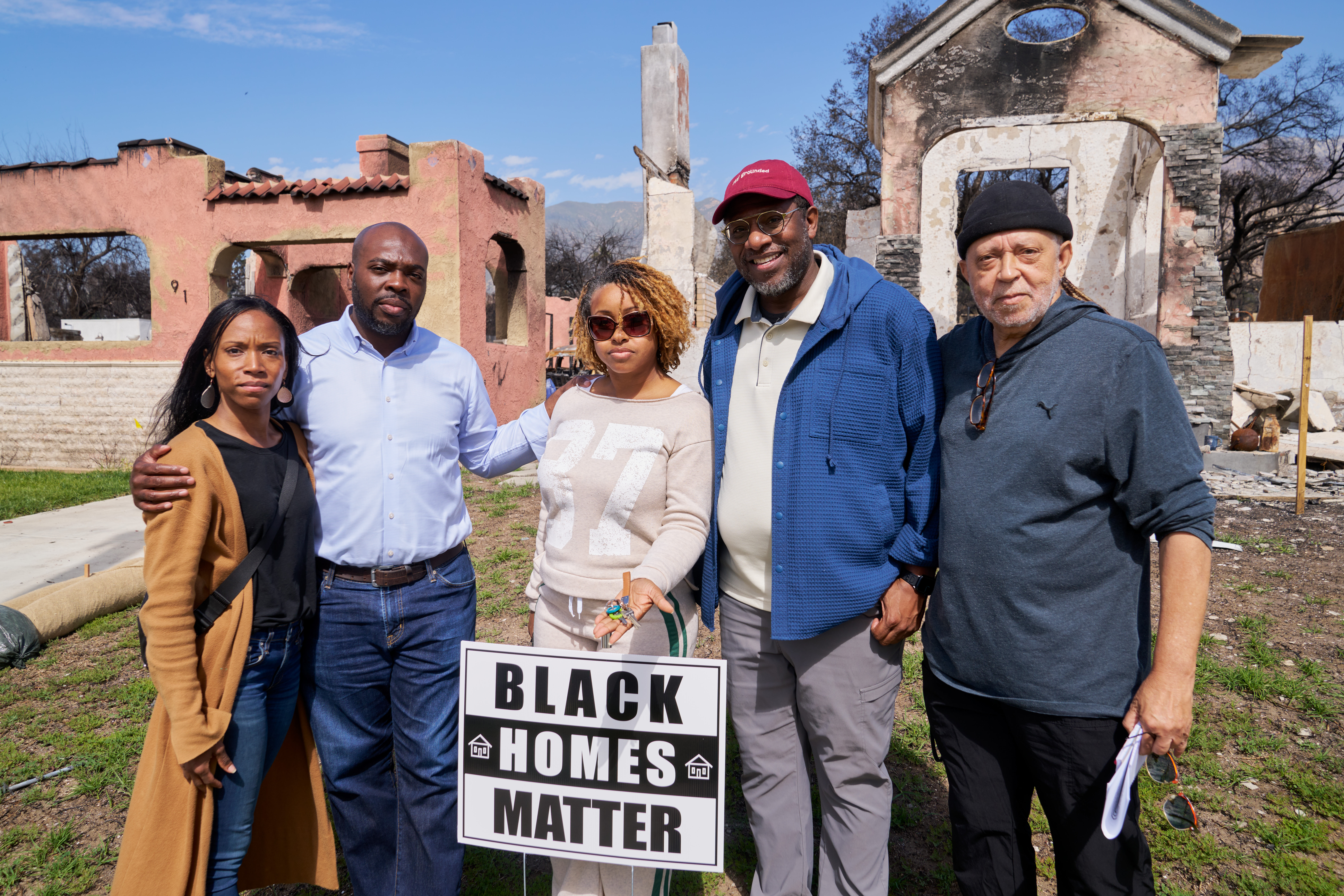Group of people standing in front of fire damaged homes with a sign that reads Black Homes Matter
