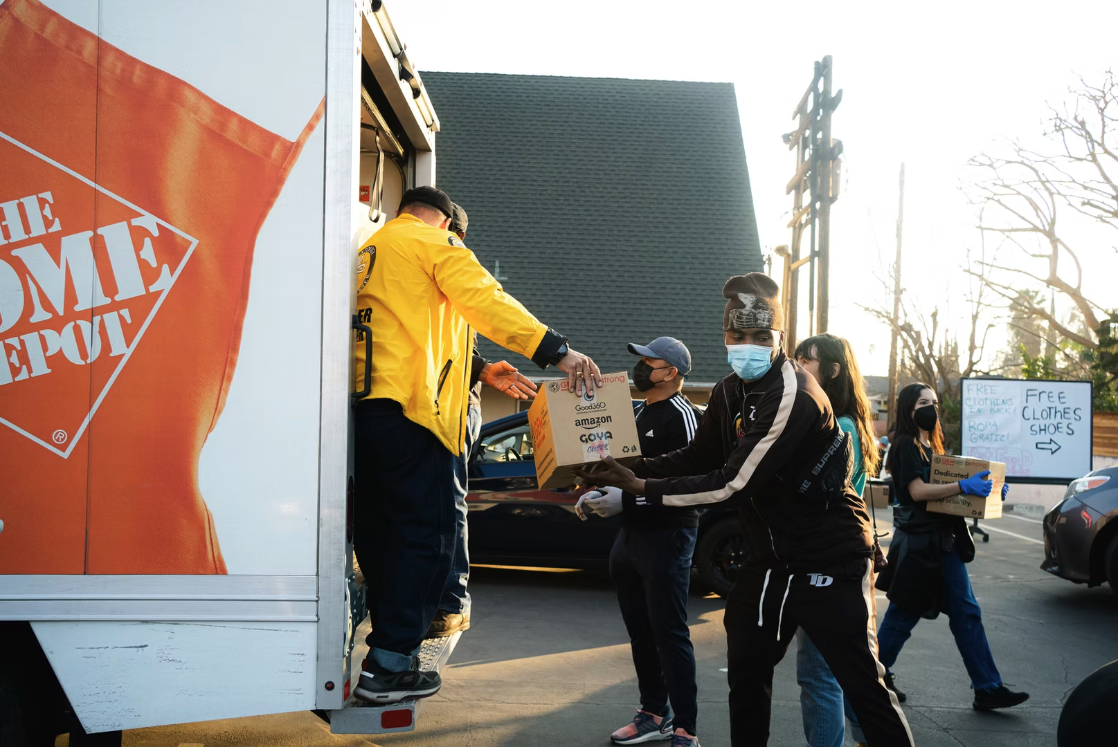 Image of people wearing masks unloading boxes from a truck in front of a sign that reads 'Free Clothes Shoes'